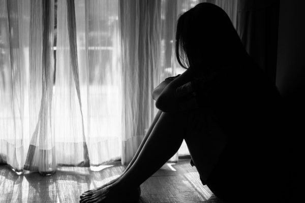 Black and white silhouette of a woman sitting on the floor in front of a window hugging her knee.
