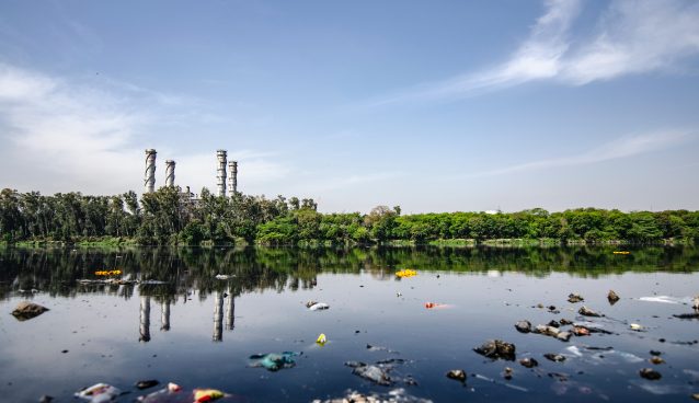 Garbage floating on a body water in front of trees and a factory.