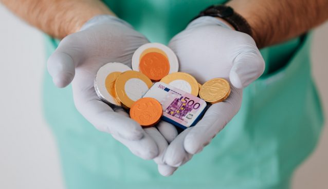 A medical professional in scrubs and gloves holds a handful of British coins.