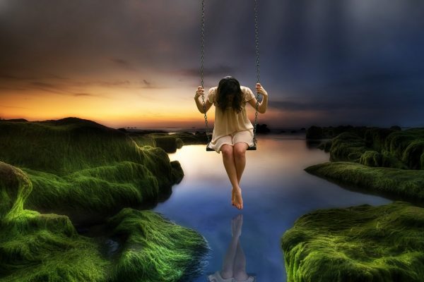 A girl sits on a swing looking down at the water below.